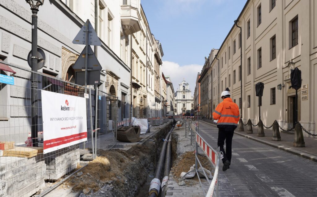 Pracownik w pomarańczowej kamizelce odblaskowej przechodzi obok wykopu na ulicy w Krakowie.